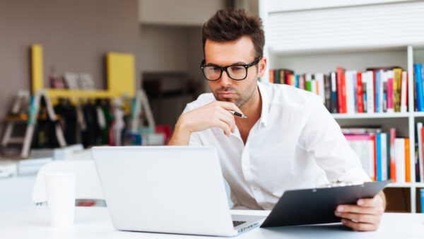 Man looks thoughtful whilst looking at laptop, with clipboard in hand